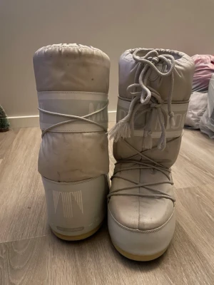 Vita Moon Boot snow boots - Moon Boot snow boots i ljusgrått med klassisk logga runt skaftet. Skorna har snörning framtill och är tillverkade i syntetmaterial med vadderad insida. Perfekta för vintern och håller fötterna varma under kalla dagar. Går att tvätta för finare färg (passar mig som jag 39 i vanliga skor)