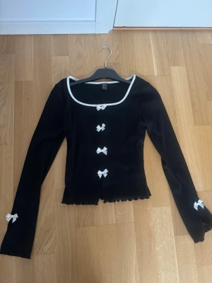 Svart kofta med vita rosetter från SHEIN - Supersöt svart kofta från SHEIN i storlek XS, med vita rosetter längs knäppningen och på ärmarna. Koftan har vit kant runt halsen, långärmad design och lätt ribbad struktur. Perfekt för dig som gillar gulliga detaljer och vill sticka ut lite extra.