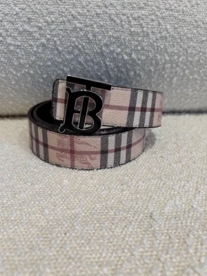  Burberry-bälte - Snyggt Burberry bälte i skinn med klassiskt rutigt mönster i beige, svart och rött på ena sidan och mörkt rutigt mönster på andra sidan. Bältet har en stor metallspänne med Burberrys ikoniska TB-logga. Perfekt accessoar för att lyfta din outfit. Man kan vända på det så att det blir 2 färger, svart och beige , kom dm för eventuella frågor 🧐 pris kan också diskuteras 💯.