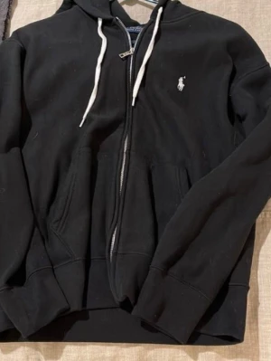 Svart Polo Ralph Lauren zip - Säljer en svart zip-hoodie från Polo Ralph Lauren. Klassisk och clean design med vit dragkedja, snören i huvan och diskret broderad Polo-logga på bröstet. Mjuk och skön passform – perfekt till vardags eller chill. En tidlös hoodie som funkar till allt.