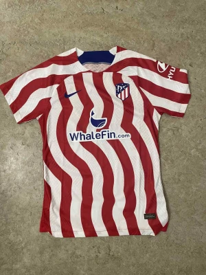 Randig Atlético Madrid Nike tröja - Säljer en officiell Atlético Madrid matchtröja från Nike med röda och vita vågiga ränder, klubbmärke på bröstet och sponsorer tryckta fram och bak. Tröjan har korta ärmar och är tillverkad i lätt polyester som andas. Perfekt för fotbollsfans!