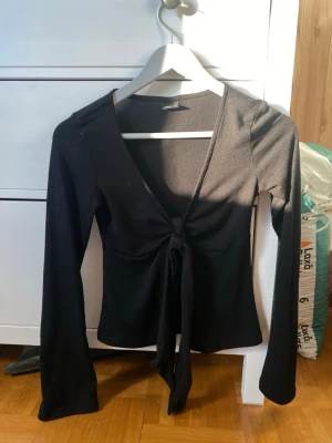 Svart topp med knyt - Säljer en svart topp från Gina Tricot med långa ärmar och snygg knytning framtill. Toppen har v-ringad hals och är tillverkad i mjuk bomullsmix som sitter skönt mot kroppen. Perfekt att styla med jeans eller kjol för en cool look.