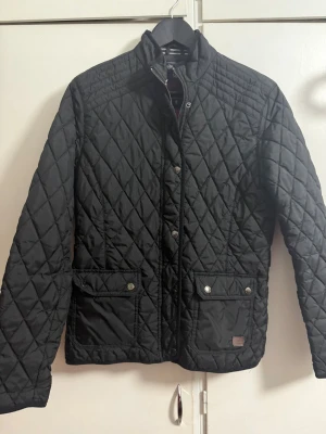 Svart quiltad jacka från South West - Quiltad svart jacka från South West med snyggt rutmönster, två stora fickor med tryckknappar och dragkedja framtill. Insidan är fodrad i vinrött och jackan har ståkrage. Perfekt för dig som gillar stilren och funktionell design.