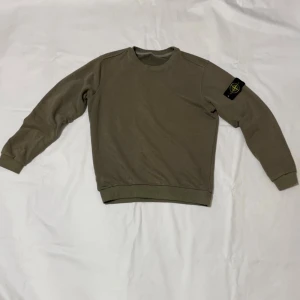 Olivgrön sweatshirt från Stone Island - Säljer en olivgrön sweatshirt från Stone Island med klassisk patch på vänster ärm. Tröjan har rund halsringning, ribbade muddar och är långärmad. Perfekt för dig som gillar stilrena och avslappnade plagg.