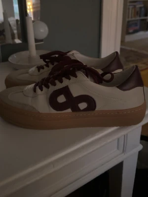 Pavement sneakers i vinrött och beige - Sneakers från Pavement med vinröda detaljer och snörning. Skorna har en beige ovandel i skinn och mocka, vinröda snören och logga på sidan samt en brun gummisula. Endast använda 1 gång, köpta för 1800 kr.