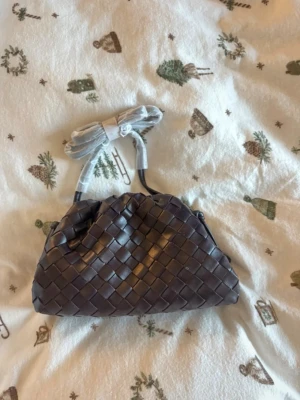 Brun flätad handväska i skinn - Säljer en snygg brun handväska med flätad design och mjuk struktur. Väskan är tillverkad i skinn och har ett rymligt innerfack med tygfoder. Medföljande axelrem gör den enkel att bära både i handen och över axeln. Perfekt för dig som gillar stilrena detaljer och klassisk look.