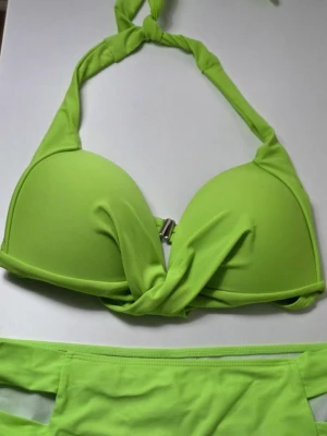 Neongrön Bikini Set Knuten Halterneck Topp | Stl L | Nyskick - En fantastiskt snygg och iögonfallande neongrön bikini i storlek L. Perfekt för stranddagar och poolhäng när du vill synas!  Överdel: Knuten halterneck-modell. Vadderade kupor med en modern vriden/ veckad design framtill. Stängs med ett spänne i ryggen.  Underdel: Klassisk bikinimodell med snygga cut-out/ banddetaljer på sidorna. Storlek: L. Skick: Helt ny och aldrig använd (endast tvättad). Säljes då den tyvärr var för stor för mig