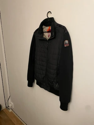 Svart quiltad jacka från Parajumpers - Säljer en svart quiltad jacka från Parajumpers med snygg patch på ärmen. Jackan har dragkedja framtill, två sidofickor med dragkedja och ribbade muddar. Materialmix med vadderad front och mjuka ärmar i fleece. Perfekt för dig som vill ha en clean och sportig look.
