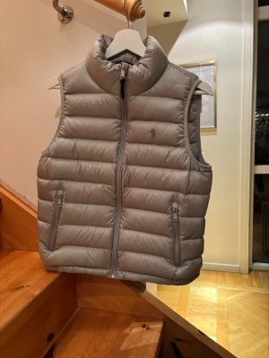 Grå dunväst från Polo Ralph Lauren - Säljer en grå dunväst från Polo Ralph Lauren. Perfekt att slänga över en hoodie eller tröja. Passar 155-165 cm.