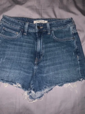 Blå jeansshorts med fransig kant - Snygga blå jeansshorts från Mimi Jeans med rå, fransig kant nertill och klassisk femficksdesign. Shortsen har normal passform och stängs med knapp och dragkedja. Perfekta för varma dagar och enkel att matcha med allt. Köptes utomlands.