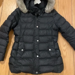 Svart dunjacka Tommy Hilfiger L - Svart dunjacka från Tommy Hilfiger med pälskantad huva och quiltad design. Jackan har tryckknappar, dragkedja och två fickor framtill. Insidan har snygga Hilfiger-detaljer med röd och vit kant. Perfekt för kalla dagar.