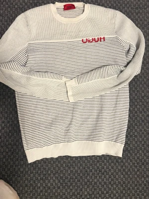 Vit randig tröja från Hugo Boss - Säljer en vit långärmad tröja från Hugo Boss med tunna svarta ränder och röd logotyp på bröstet. Tröjan har rund hals och är stickad i bomull, vilket gör den både mjuk och skön. Perfekt för dig som gillar stilrena och klassiska plagg med en modern touch.