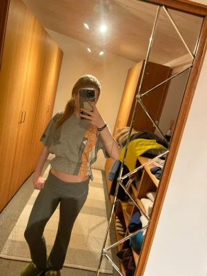 Grå croppad t-shirt från en collab med Adidas och Stella mcartny - Säljer en grå croppad t-shirt från en collab med Adidas och Stella Mcartny med orange och grått mönster framtill. Toppen har korta ärmar, resår i midjan och tryckdetaljer på både bröstet och ärmen. Perfekt för en sportig och avslappnad stil.