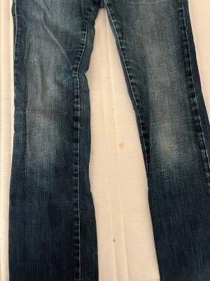 Mörkblå jeans med knappficka - Snygga mörkblå jeans med slitningar och en unik bakficka med knappdetalj. Jeansen har klassisk rak passform och är tillverkade i robust denim. Perfekt för dig som gillar en cool och avslappnad stil med lite extra detaljer på fickan.