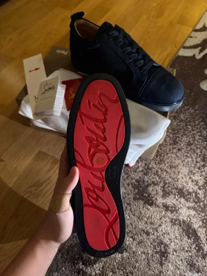 Marinblåa sneakers från Christian Louboutin - Säljer ett par marinblåa sneakers från Christian Louboutin med klassisk röd sula och snygg snörning. Skorna är tillverkade i mocka och har en rund tå samt platt sula. Märkets logga syns på både sula och häl. Perfekt för dig som gillar exklusiva detaljer.