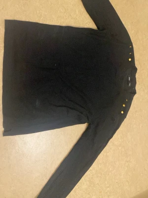 Svart stickad tröja med guldknappar Sinsay - Svart stickad tröja från Sinsay i storlek M. Tröjan har långa ärmar och tre guldknappar på ena axeln som detalj. Klassisk rund halsringning och mjukt material som passar perfekt till höst och vinter.