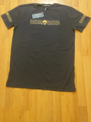 Svart  t-shirt med gulddetaljer - Svart t-shirt  i bomull med guldbroderade grekiska mönster och Medusa-logga på bröstet. Kortärmad modell med gulddetaljer även på ärmarna och en liten tag på sidan. Snygg och lyxig vibe för dig som gillar statement-plagg.