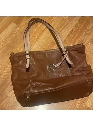 Brun handväska från Michael Kors - Snygg handväska i brunt skinn från Michael Kors med ljusa handtag och metallfästen. Väskan har MK-logga i silver och praktisk dragkedja upptill. Perfekt storlek för att få plats med allt du behöver och stilren design som lyfter din look. Skick se på bilderna sliten på vissa ställen och lite smutsig inuti (har inte försökt tvätta bort) men inget störande men äkta