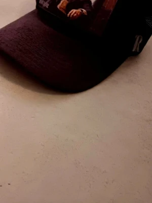 Svart meshkeps från Rovior - Säljer en svart keps från Rovior med mesh på sidorna och justerbart spänne bak. Framtill finns en tryckt bild och ett stort R broderat på sidan. Kepsen har en böjd skärm och insidan är dekorerad med Rovior-loggor. Perfekt för dig som gillar streetwear.