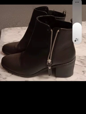 Svarta boots med dragkedja, strl 39 - Säljer ett par svarta boots i skinn med grov dragkedja på sidan och rund tå. Skorna har en låg klack och stilren design som passar till många olika outfits. Perfekt för dig som gillar klassiska och snygga skor med lite edge. Använd bara 1 gång.