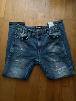 Blå jeans med slitningar - Snygga blå jeans från Icono med coola slitningar och tvättade detaljer framtill. Klassisk femficksmodell med bälteshällor och knappgylf. Jeansen har en rak passform och är tillverkade i jeansmaterial med en något mörkare blå ton. Skriv för mer bilder frågor. Pris kan diskuteras 