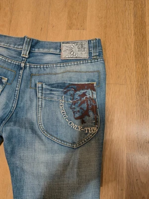 Diesel blå jeans med tryck bakficka - Snygga blå jeans från Diesel med coolt tryck på bakfickan och klassisk patch vid linningen. Jeansen har raka ben, normal passform och slitna detaljer för en avslappnad vibe. Fickor fram och bak, samt knappar och dragkedja i metall.