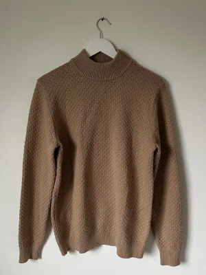 Beige polotröja från Uniqlo, M - Säljer en beige polotröja från Uniqlo i storlek M. Tröjan har ett snyggt stickat strukturmönster och ribbad krage, ärmslut och nederkant. Materialet är 60% bomull och 40% akryl, vilket gör den både mjuk och skön. Perfekt för dig som gillar enkel och stilren design. 5L2