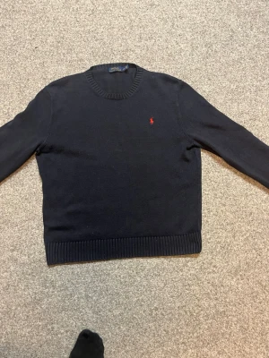 Mörkblå stickad tröja Polo Ralph Lauren - Äkta. Snygg mörkblå stickad tröja från Polo Ralph Lauren med klassisk röd broderad logga på bröstet. Rund halsringning och ribbade muddar vid ärmslut och nederkant. Perfekt för dig som gillar stilrena och tidlösa plagg.