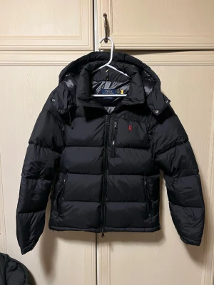 Svart pufferjacka från Polo Ralph Lauren - Svart pufferjacka från Polo Ralph Lauren med huva och dragkedja framtill. Jackan har det klassiska röda Polo-logot broderat på bröstet och två sidofickor med dragkedja. Perfekt för kalla dagar och har en clean, sportig look. Nypris: 5995kr. Aldrig använd, endast testad. Helt felfri och fläckfri. Fraktar snabbt så den hinner komma innan jul.