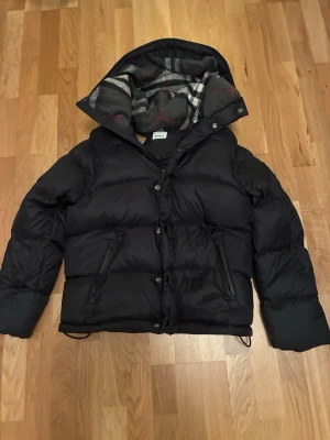 Svart pufferjacka från Burberry - Svart pufferjacka från Burberry med klassisk logopatch på ärmen och rutigt foder i huvan. Jackan har dragkedjor på fickorna, knappar framtill och långärmade avtagbara ärmar. Perfekt för kalla dagar och har en riktigt snygg streetstil.