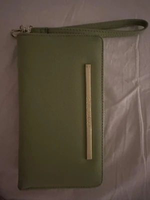 Grön plånbok från Steve Madden - Snygg grön plånbok från Steve Madden med guldfärgad metall-detalj på framsidan och matchande dragkedja. Utsidan är stilren och insidan har flera fack för kort, mynt och ID-fönster. Handledsrem gör den enkel att bära med sig. Pris kan diskuteras.