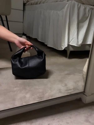 Svart handväska med gulddetalj - Snygg svart handväska i skinnimitation med en chunky guldfärgad detalj på handtaget. Väskan har en mjuk, boxig form och medföljer en extra axelrem. Perfekt storlek för att få plats med allt nödvändigt och passar till många olika stilar.