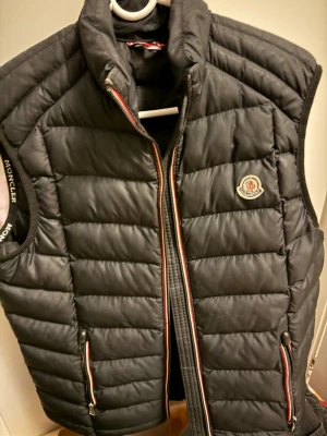 Svart dunväst från Moncler - Säljer en svart dunväst från Moncler med quiltad design och klassisk logotyp på bröstet. Västen har dragkedja framtill med röd, vit och blå detalj samt två fickor med matchande dragkedjor. Perfekt för lager-på-lager under kyliga dagar.