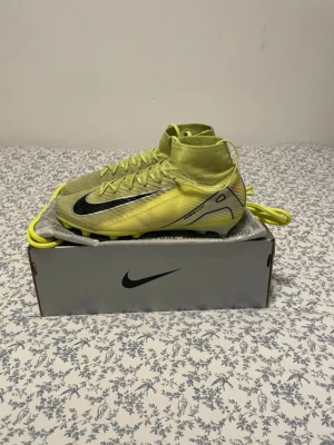 Nike Mercurial Superfly Elite AG - Säljer ett par grymma Nike fotbollsskor i ljusgul färg med svart swoosh och coola grafiska detaljer på sidan. Skorna har snörning, platt sula med dobbar och en strumpliknande hög krage för extra stöd. Materialet är syntet och designen är riktigt modern.