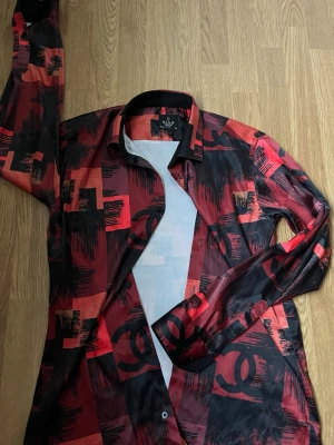 Röd och svart slim fit skjorta Hang London - Snygg slim fit skjorta från Hang London i storlek M. Skjortan har ett unikt röd-svart mönster med grafiska rutor och penseldrag, samt knappar framtill. Materialet känns lätt och glansigt, perfekt för dig som gillar att sticka ut med färg och design.