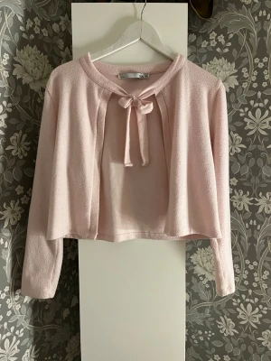 Ljusrosa kofta med knyt - Supersöt ljusrosa kofta med knyt framtill och lång ärm. Koftan är croppad och har en mjuk, lätt textur som gör den perfekt att slänga över en topp. Lite smått nopprig men inget man tänker på!