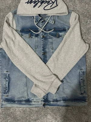 FSBN jeansjacka med hoodie XXL - Snygg jeansjacka från FSBN i storlek XXL med ljusgrå sweatshirt-ärmar och huva. Jackan har klassisk blå denimfront med knappar och två bröstfickor. Huva med broderad text och dragsko för extra streetstyle-vibe.