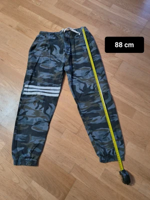 Camo cargopants med snörning och gummiband  - Säljer ett par camouflagemönstrade cargopants med elastisk midja och snörning. Byxorna har en bakficka och resår vid bensluten. Färgerna går i ljusblå, brun och svart.  och byxorna har vita ränder på ena benet.