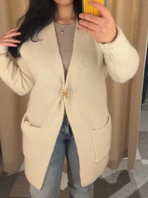 Beige stickad kofta - Mysig beige stickad kofta med lång ärm och två stora fickor framtill. Perfekt att slänga över en t-shirt eller topp för en chill och stilren look.