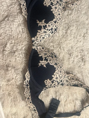 Mörkblå spetsdetaljerad väst från intimissimi  - Säljer en mörkblå väst med eleganta spetsdetaljer i beige längs kanterna. Västen har en feminin form och är tillverkad i ett mjukt syntetmaterial. Spetsen har ett blommigt mönster som ger en lyxig känsla. Perfekt för dig som gillar stilrena och romantiska plagg.