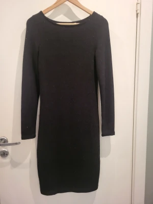 Svart glittrig långärmad klänning - En enkel och stilren svart glittrig klänning med lång ärm och rund hals. Klänningen har en rak passform och är knälång, perfekt för dig som gillar minimalistisk design och vill ha något bekvämt men snyggt på festen.