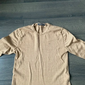 Beige stickad tröja från J.Lindeberg - Snygg beige stickad tröja från J.Lindeberg med rund halsringning och diskret broderad logga på bröstet. Tröjan har lång ärm och ribbade muddar vid ärmslut och nederkant. Perfekt för dig som gillar stilrena och klassiska plagg. Använd ca 1 gång eftersom den var för liten.