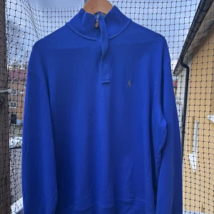 Blå half zip tröja från Ralph Lauren - Snygg blå långärmad tröja från Ralph Lauren med half zip och läderdetalj på dragkedjan. Tröjan har ett broderat RL-märke på bröstet och är gjord i mjuk bomull. Perfekt för dig som gillar klassisk stil med en modern touch.Priset är ristat i sten går ej att pruta!!