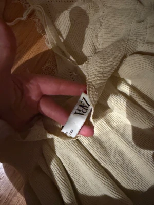 Beige stickad linnetopp från Zara - Säljer en beige stickad linnetopp från Zara med smala axelband och v-ringad hals. Toppen har fina spetsdetaljer vid urringningen och ett ribbat mönster över hela plagget. Den är luftig och har en lätt volang nedtill för extra flow.