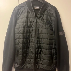 Barbour cardigan/hybrid - Svart quiltad bomberjacka från Barbour International i storlek M. Jackan har ribbad krage, muddar och dragkedja framtill. Materialet är bomull med stickade ärmar och quiltad front. Snygg patch på ärmen och två fickor med dragkedja.