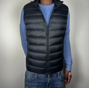 Uniqlo väst - Stilren svart dunväst med horisontella sömmar och hög krage. Västen har en klassisk passform och stängs med dragkedja framtill. Perfekt att slänga över en hoodie eller tröja för extra värme. Enkel och clean look som funkar till det mesta.