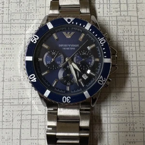 Emporio Armani blå-silver klocka - Stilren klocka från Emporio Armani med blå urtavla och silverfärgad länk i metall. Klockan har kronograf, tydliga index och vridbar blå bezel. Snygg och sportig design med robust känsla och märkets logga på urtavlan.