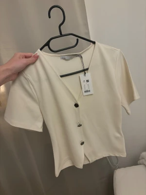 Krämvit blus med v-ringning från Lager 157 - Säljer en stilren krämvit blus från Lager 157 i storlek XS. Toppen har v-ringning och tre dekorativa knappar framtill. Materialet är mjukt och känns skönt mot huden. Perfekt för dig som gillar minimalistisk och clean stil. Helt oanvänd och köpes för 150kr säljs för 80kr (pris sänks inte) 