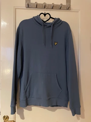 Blå hoodie från Lyle & Scott - Snygg blå hoodie från Lyle & Scott med klassisk gul fågel-logga på bröstet. Tröjan har huva med snörning, stor magficka och ribbade muddar. Använd men i bra skick och säljs i befintlig skick. 