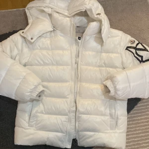 Vit Moncler pufferjacka med huva - Säljer en clean vit pufferjacka från Moncler i storlek M. Jackan har en stor svart Moncler-logga på ärmen, huva och dragkedja framtill. Fylld med dun och har två sidofickor. Perfekt för kalla dagar när du vill ha både stil och värme. Priset kan diskuteras!